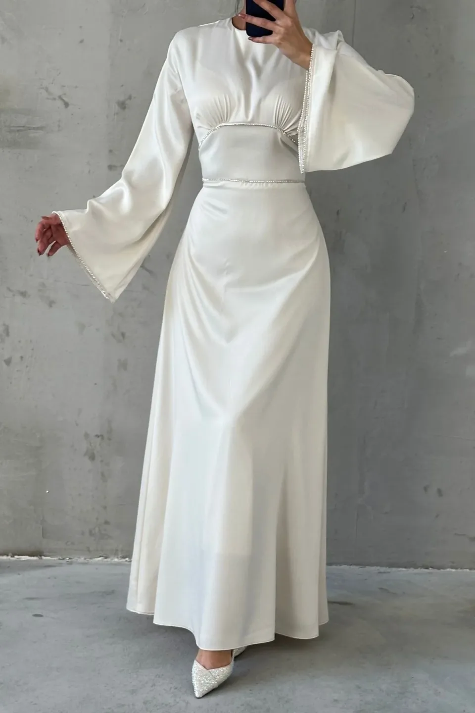 White maxi dress with wide, flowing sleeves and contrasting white trim along the neckline, waist, and cuffs. The dress is made of a smooth, draping fabric.