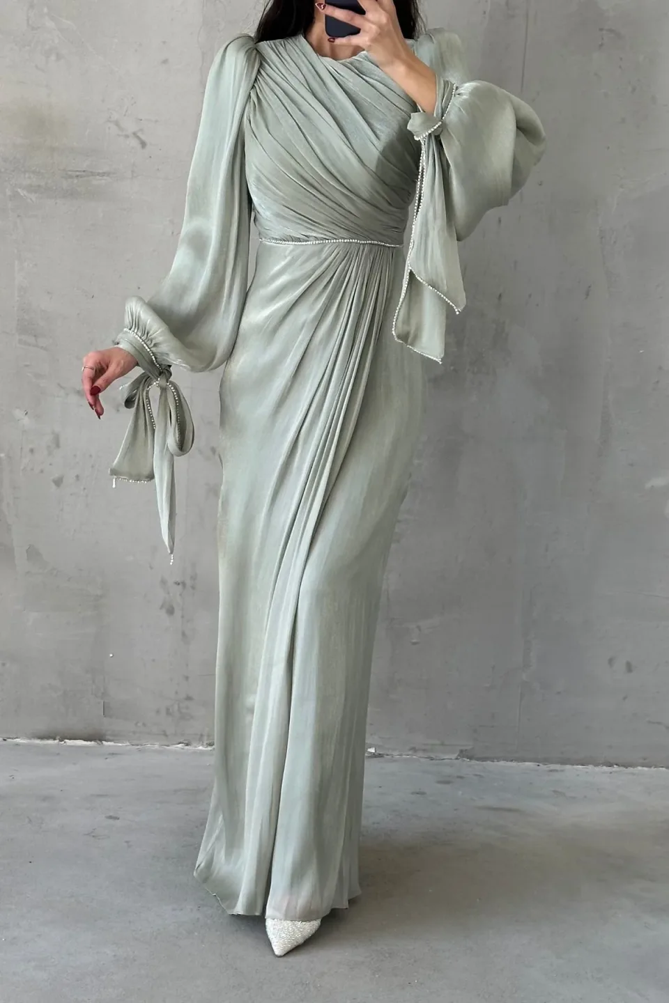 A long, dark brown maxi dress with draped detailing and sparkling trim, displayed against a textured gray background.