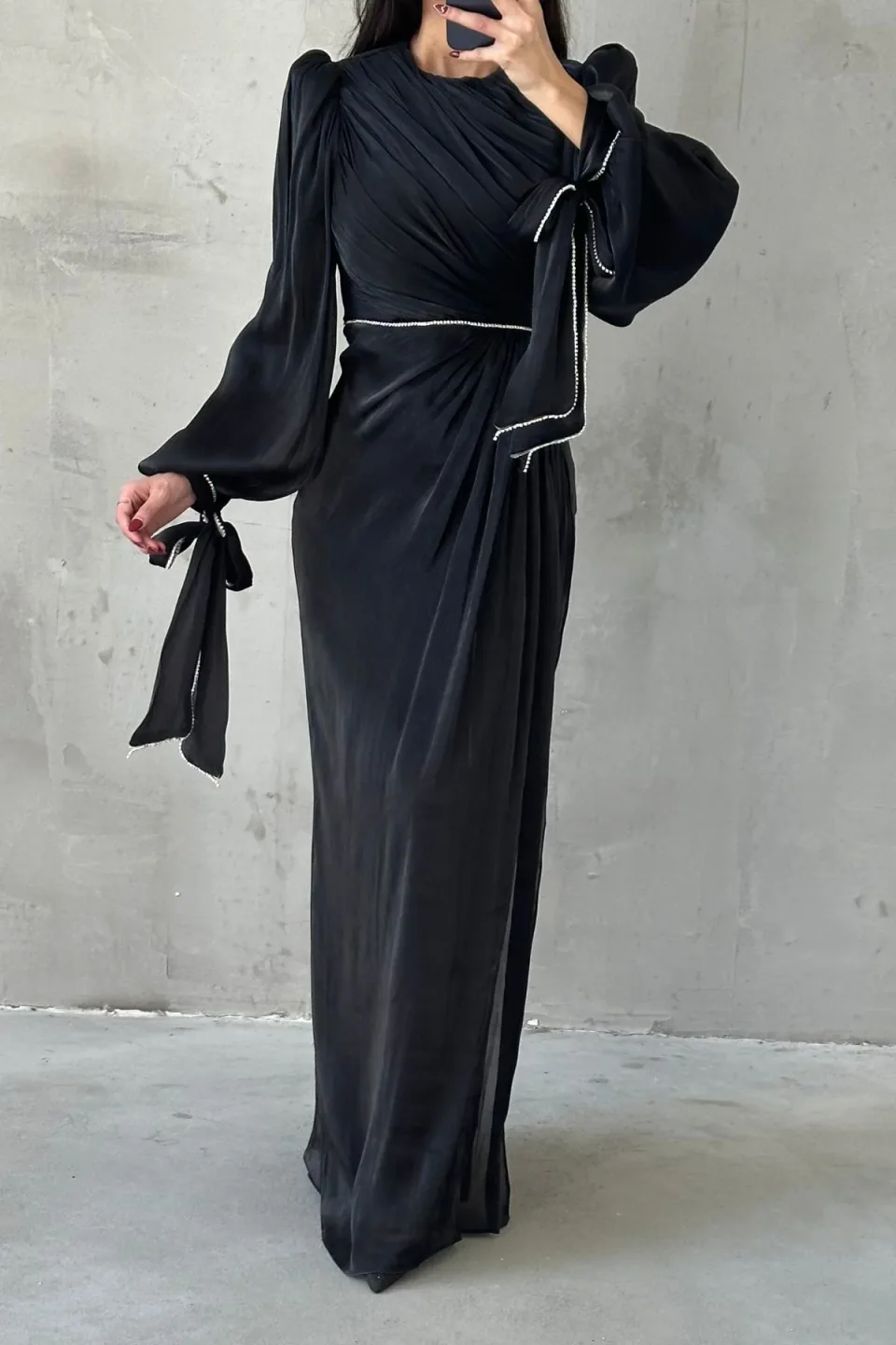 A long, black maxi dress with draped detailing and sparkling trim, displayed against a textured gray background.