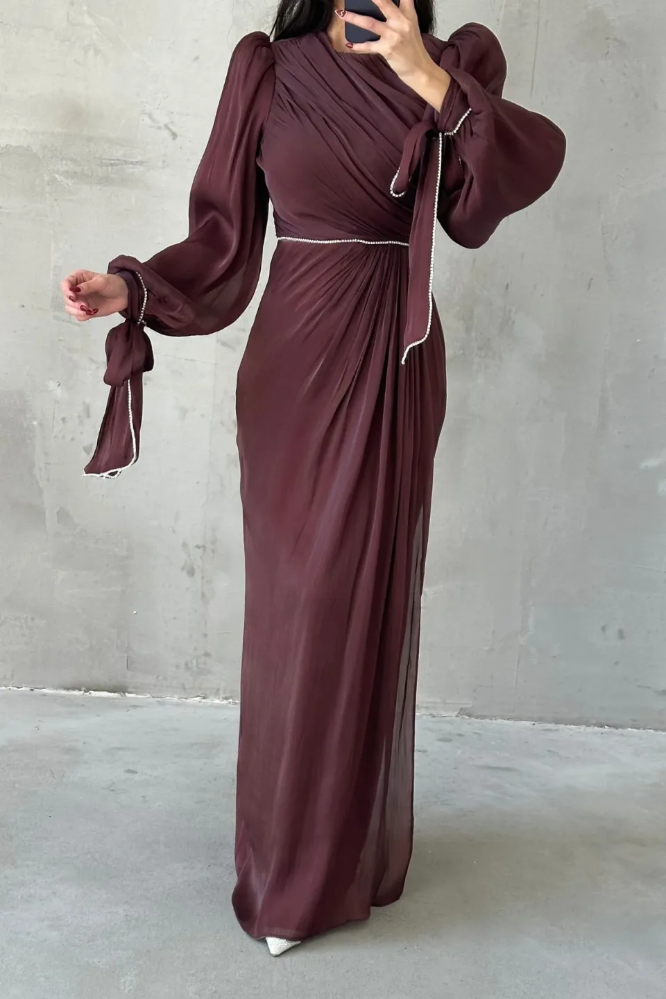 A long, dark brown maxi dress with draped detailing and sparkling trim, displayed against a textured gray background.