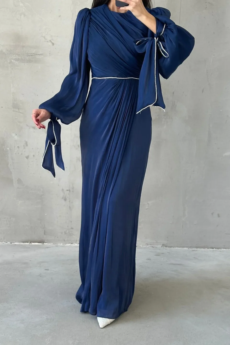 A long, navy blue maxi dress with draped detailing and sparkling trim, displayed against a textured gray background.