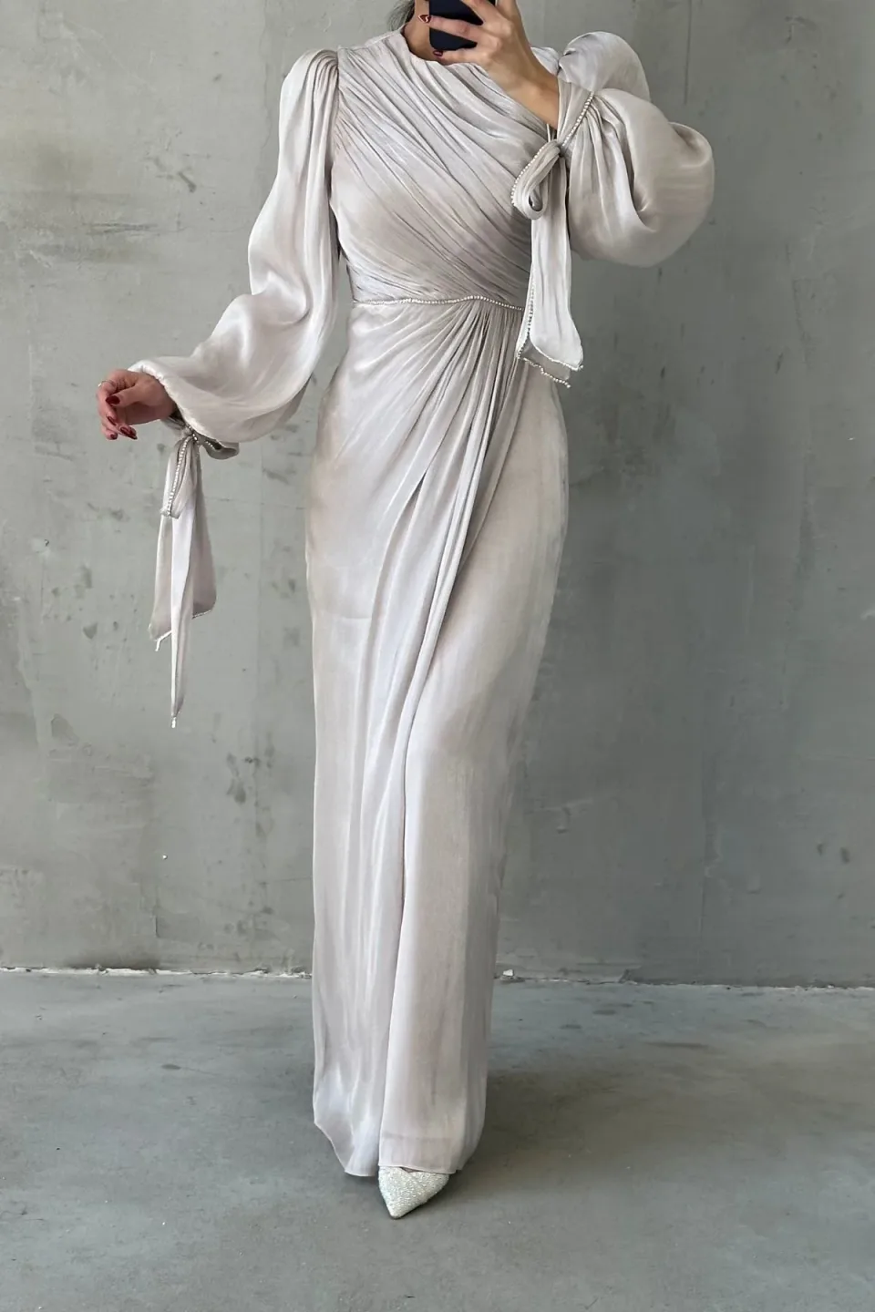 A long, silver maxi dress with draped detailing and sparkling trim, displayed against a textured gray background.