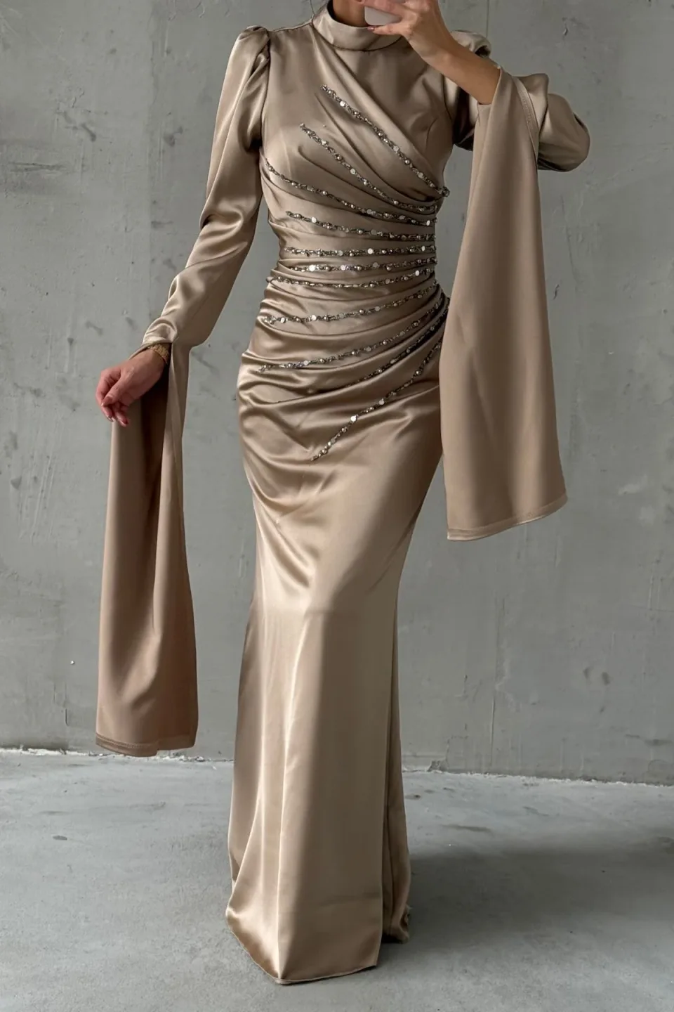 This sophisticated long-sleeved gown features a sleek, figure-hugging silhouette, a high neckline, and a beautifully embellished bodice, culminating in dramatic, flowing fabric panels that cascade from the sleeves.