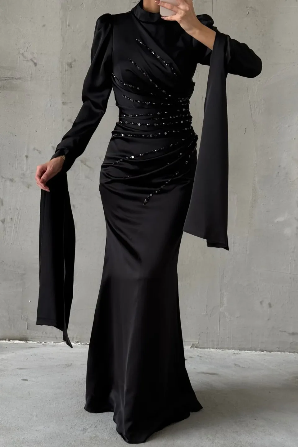 This sophisticated long-sleeved gown features a sleek, figure-hugging silhouette, a high neckline, and a beautifully embellished bodice, culminating in dramatic, flowing fabric panels that cascade from the sleeves.